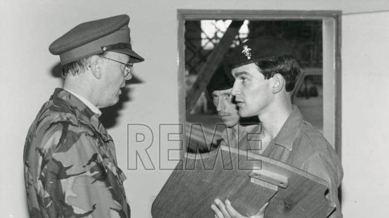 A man in military uniform speaks to two more men in uniform. One holds a sporting shield. Window behind them in background.