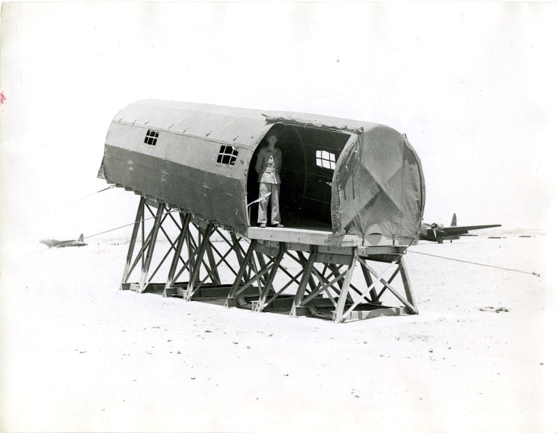 Black and white photo of a mock body of a plane on stilts. A man stands inside and is seen through a cut out on the side.