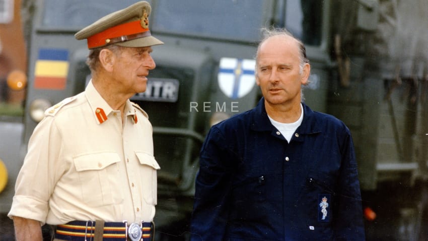 Prince Phillip in military uniform and Sergeant Jones talking. Military vehicle out of focus in background.  
