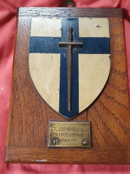 Plaque with a shield painted white with a blue cross and grey sword. A metal rectangle underneath the shield reads "CPL Conoway D.S. France Germany 1944-45" . 