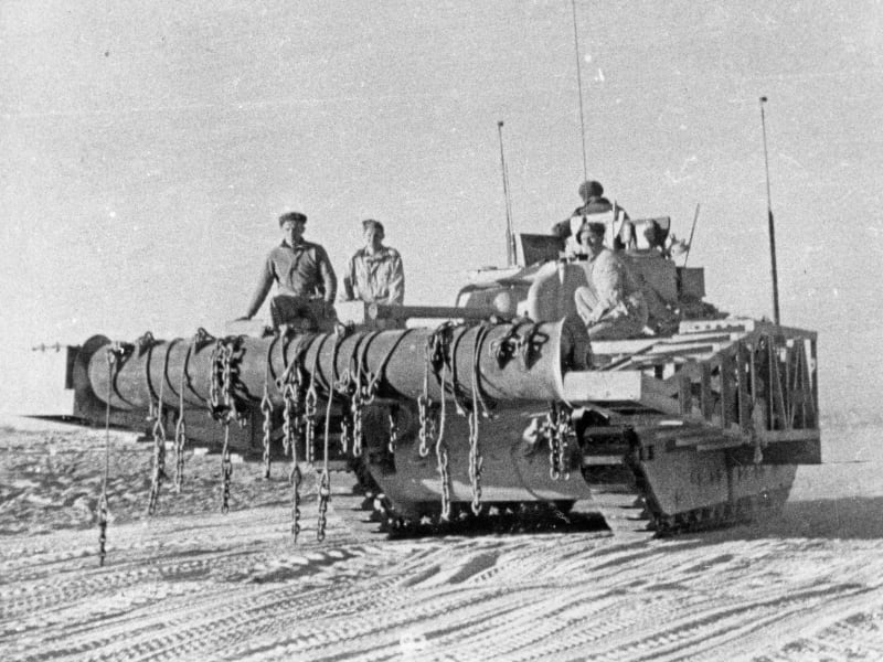 Black and white photograph of a flail tank in the desert (tank with rotator fixed to an arm out the front, chains attached to rotator), crew are sitting on the front of the tank.
