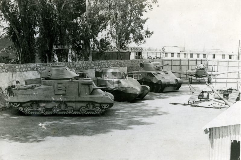Black and white image of three tanks and a small plane stationed in a walled area. Buildings and trees outside the wall in the background.