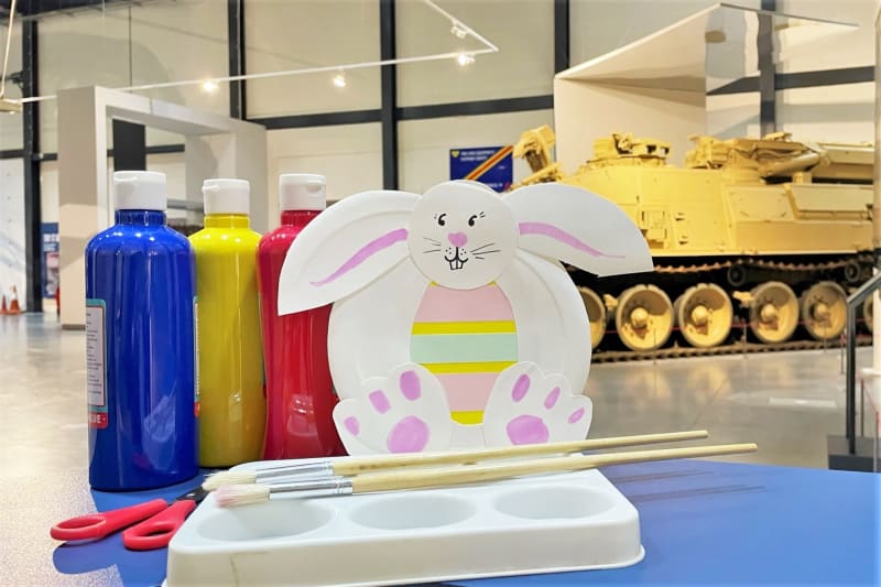 Photo of craft materials including red yellow and blue paint, an easter bunny made out of a paper plate, pictured in a vehicle gallery with tracked vehicle in the background.