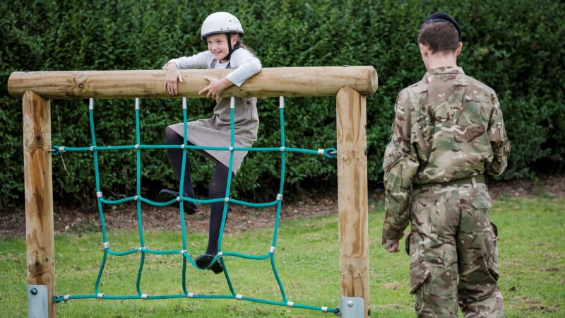 A girl wearing a helmet climbs over an obstacle, soldier in uniform watches on.