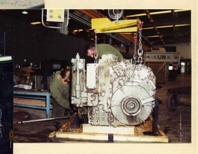 A silver coloured engine attached to a winch inside a workshop, two men in green overalls working on it.
