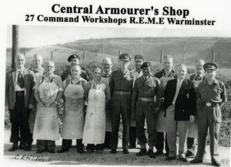 Black and white photo of a group of men outside, smiling at the camera. Some wear military uniform, some wear aprons, some wear suits. Photo caption at the top reads " Central Armourer's Shop. 27 Command Workshops R.E.M.E. Warminster ". 