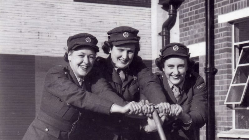 Black and white photo of three women in military uniforms holding onto a wooden handle. Brick buildings behind them.