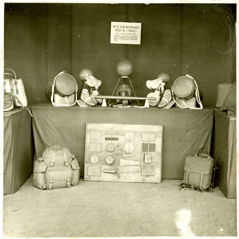 Black and white photo of a clothed table with old fashioned footballs, football boots and bags displayed on top. On the ground in front of the table are two rucksacks and a large case with materials secured inside.