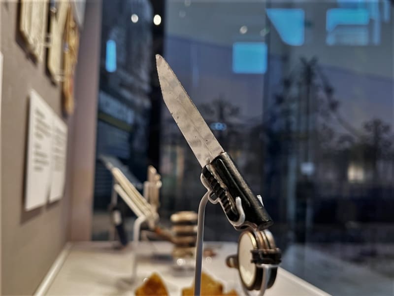 A small knife with sharp silver blade and black plastic handle, mounted on a wire mount inside a glass display case, other objects out of focus in the background.