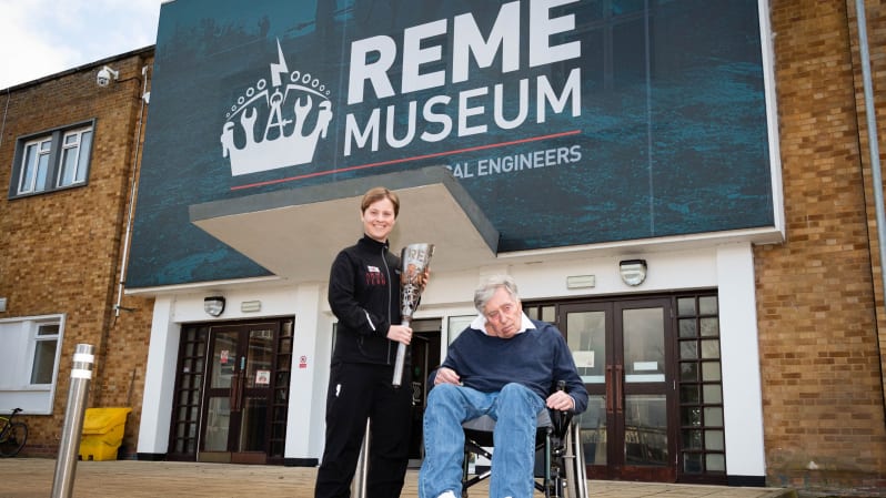 Becky Hoare holds relay torch and stands next to Jim Fox in wheelchair outside front of REME museum. Sign above entrance doors with REME logo reads: " REME Museum " .