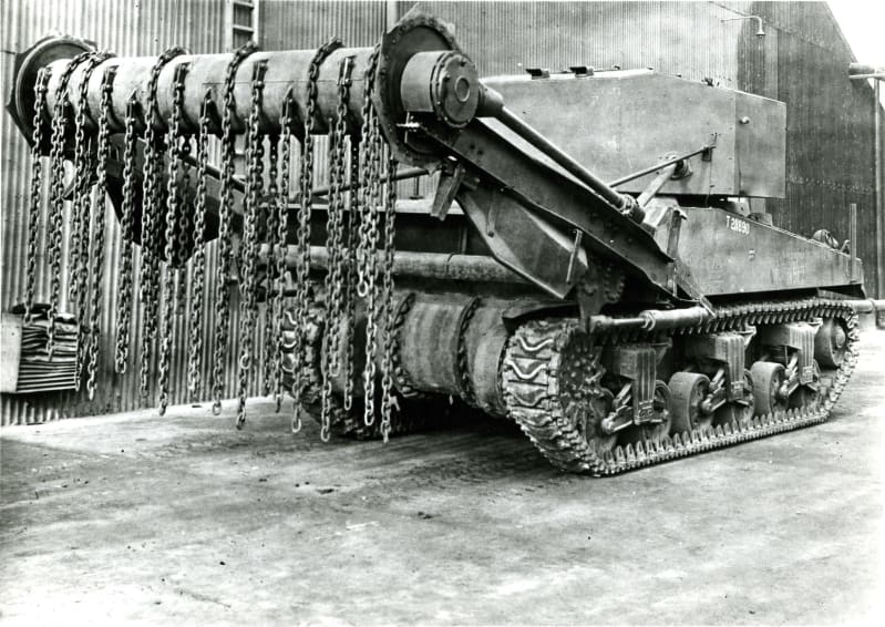 Black and white photo of a tank with long arms out front raised in the air, with roller attached, to which chains are attached and hanging down. 