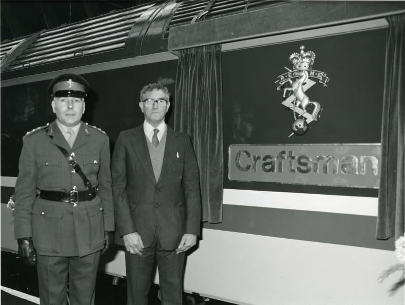 Black and white photo of a REME Officer in uniform and a gentleman in a suit standing next to a train with curtains opened to unveil a REME cap badge and name plate reading "Craftsman".