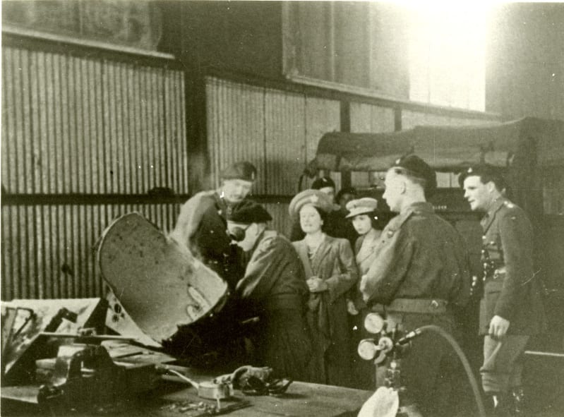 Black and white photo of the Queen Mother and Princess Elizabeth stood looking at a soldier wearing goggles working on metal, inside a workshop. Other soldiers stand around them.