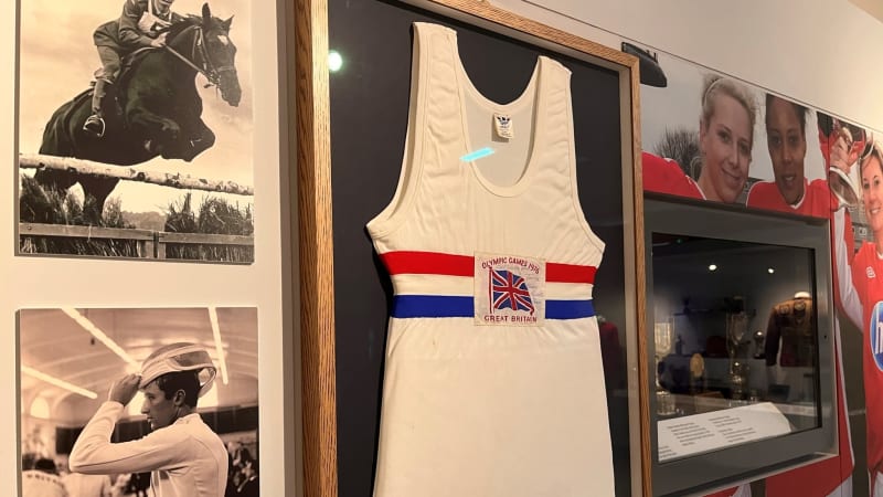 A framed vest, white with red and blue horizontal stripes across the chest, mounted on a wall with images on one side and a display case on the other.