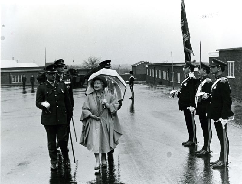 Black and white image of the Queen Mother walking and holding an umbrella under rain. Senior officers walk beside and behind, three other soldiers stand to attention to the right.