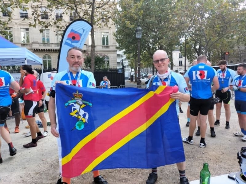 Two men in blue cycling jerseys hold the REME flag, which is blue with a red and yellow stripe running diagonally and a small logo in the top left corner. More people in ncycling jerseys in the background on the pavement.
