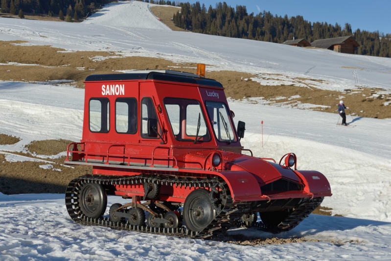 A large, red, tracked vehicle on a snow-covered hill. A person skis near the vehicle and trees are in the distant background. 