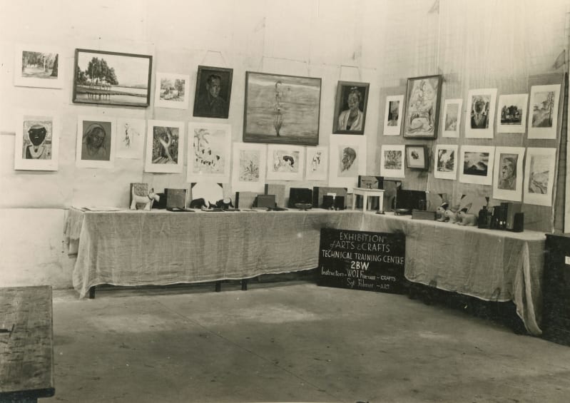 Black and white photo of an L-shaped table with models, books and other craft items on top. Paintings and sketches hung up on wall behind.