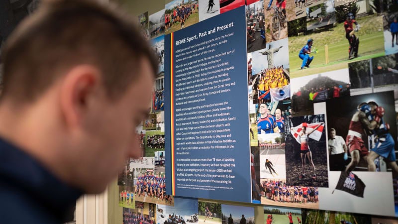 Head of man out of focus in foreground. He looks at a display of REME sport, which is pictures of athletes in various sports and a block of text with the title " REME Sport, Past and Present " 