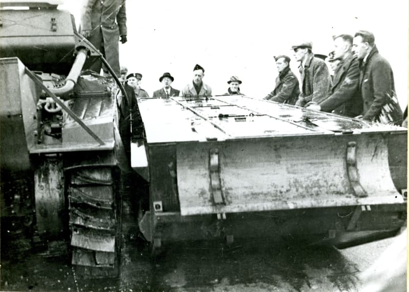 Black and white photograph of a large flat object next to a tracked vehicle, partially visible, with men stood around the side of the object.