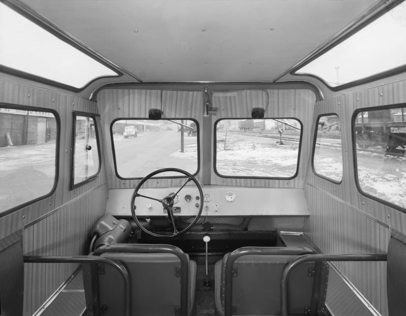 Black and white photograph of a vehicle interior, looking from the back out to the front. There are two seats, a steering wheel and six windows. The vehicle is on a road with snow on the paths. 