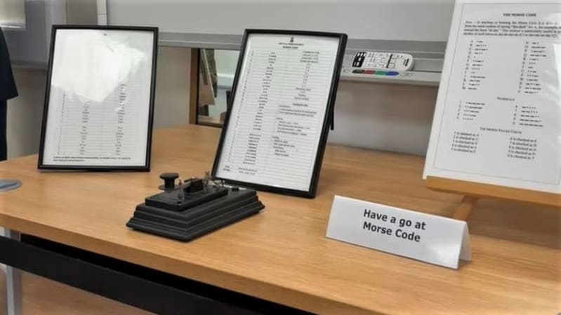 A small manual morse code machine on a table with morse code translator sheets in photo frames behind.