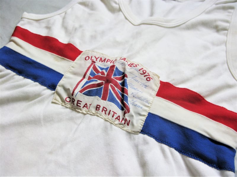 Close up of a white vest with 3 red, white and blue horizontal stripes across the middle, a white rectangular patch over the top with union flag and reads " Olympic Games 1976, Great Britain ".