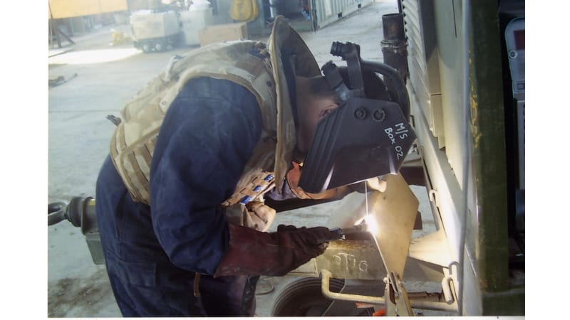 Image shows a metalsmith wearing a mask leaning over and welding a piece of metal.