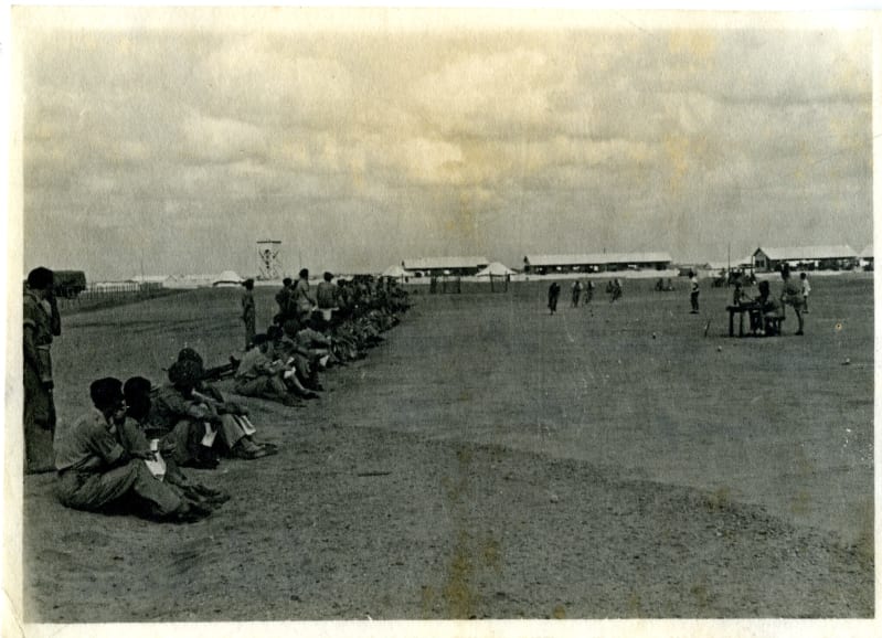 Black and white photo of a long row of soldiers sat outside. Tents and people on bikes in background.