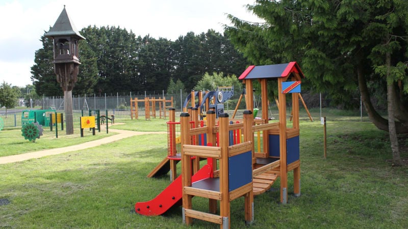 A colourful wooden park structure with a slide and bridge on some grass. Trees and more park structures in the background.