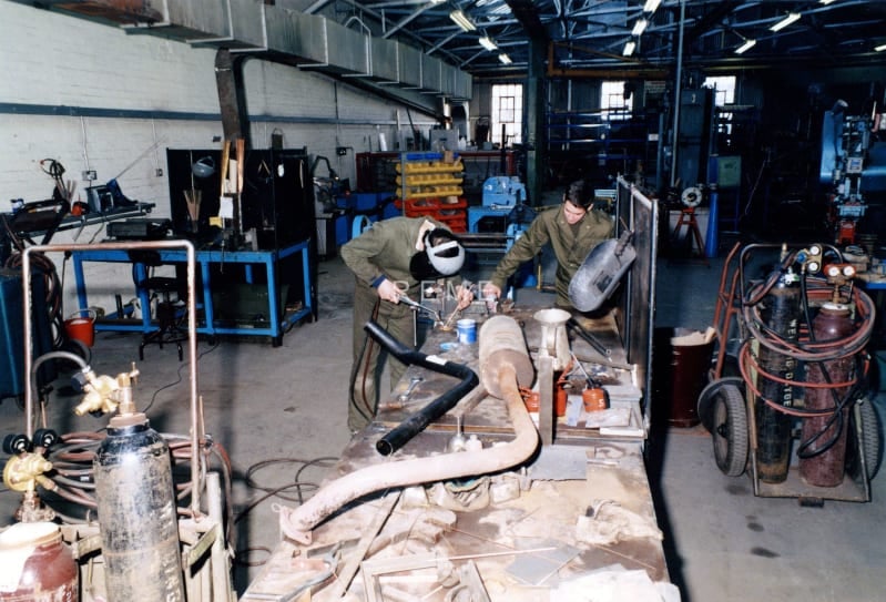 Image shows two men in green overalls working in a workshop. Tools and machinery are visible.