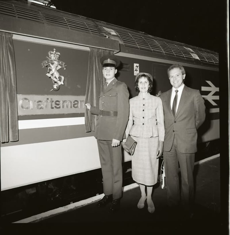 Image shows a REME soldier in uniform, a woman and a man stood next to a train with curtains opened to unveil a REME cap badge and name plate reading "Craftsman"