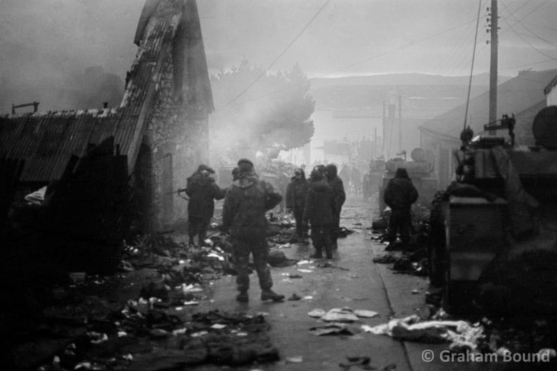 Image shows men standing around with damaged buildings and debris all over the place, foggy background