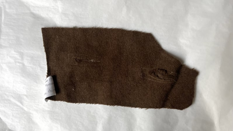 A small oblong patch of brown woven fabric with two patches sewn up in the middle, on white tissue paper.
