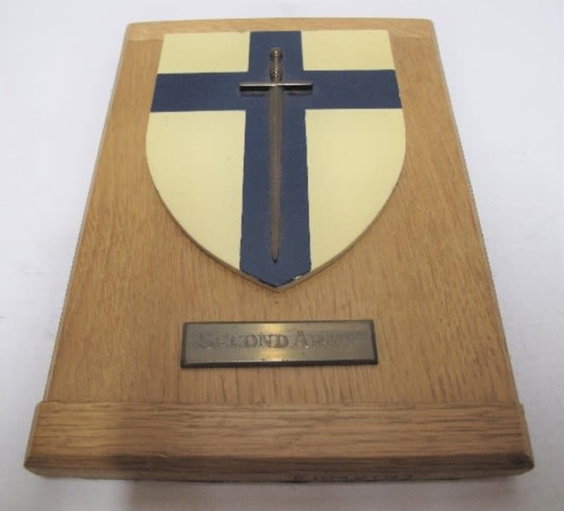 Plaque with a shield painted white with a blue cross and grey sword. A metal rectangle underneath the shield reads "Second Army" .