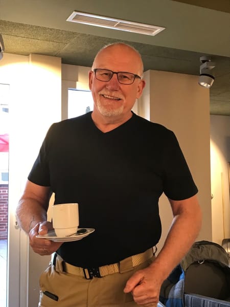 Graham in black shirt holds a saucer and teacup in one hand. Setting in restaurant.