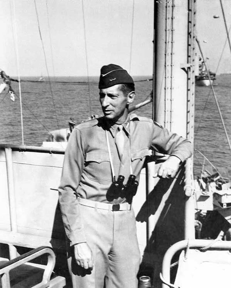 Black and white photo of a man in army uniform, binoculars hanging from his neck, stood on a ship at sea leaning against the railings. 