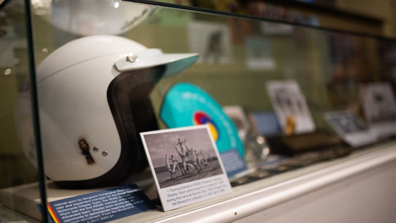 White motorcycle helmet in display case with other items out of focus in the same case. Black and white photo in front of helmet shows people performing acrobatics. 