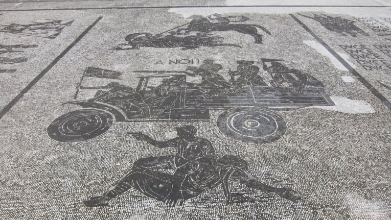 Black and white mosaic art in the style of the Romans on a pavement. Depict a soldier on horseback spearing another soldier on the ground, soldiers in an army truck, and two soldiers on the floor, one holds a gun.