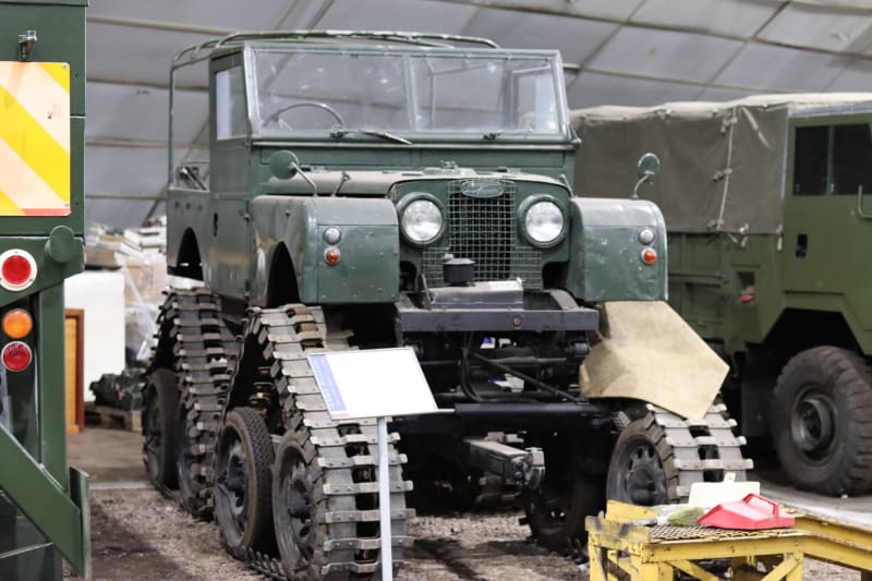 A green open top Land Rover with sets of tracked wheels where wheels normally would be. Parked in between other vehicles inside a hangar.