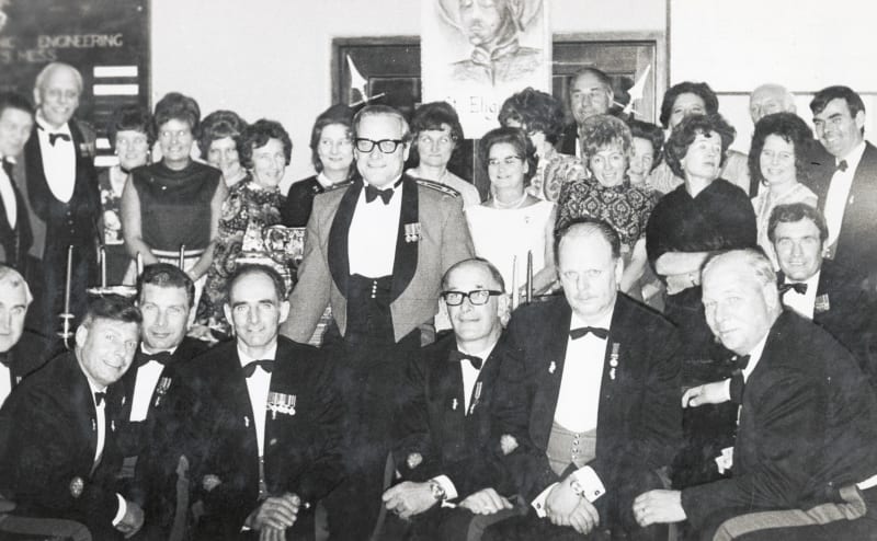 Black and white photo of room filled with people in smart dress. All look at camera.