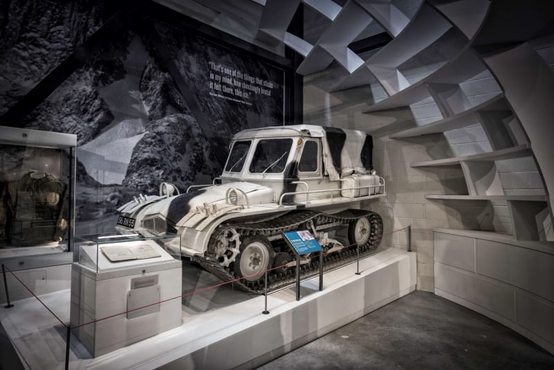 A white and black tracked vehicle sitting on a plinth inside a gallery. An igloo structure and display cases surround it.