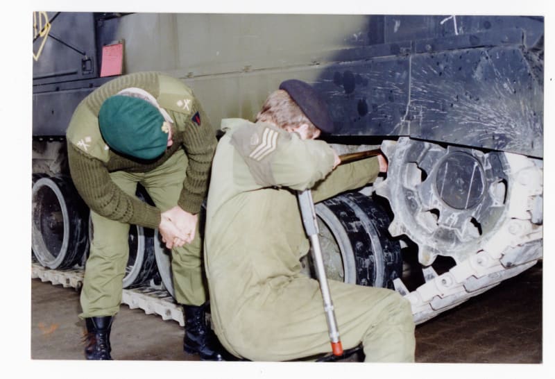 Two soldiers in green coverall working on the tracks of a large vehicle.