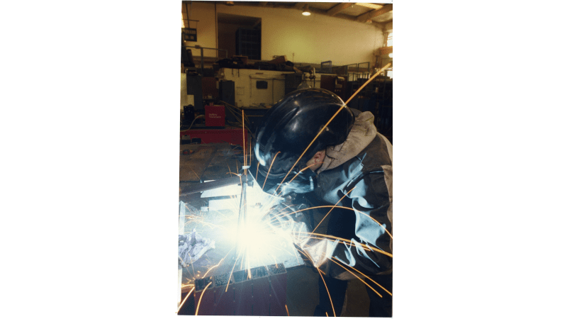 Image shows a welder leaning onto a bench, wearing a large welding mask/helmet, sparks are flying out from the metal.