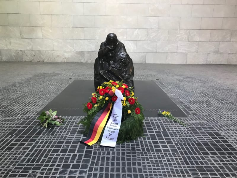 Small statue of a mother mourning her son on a path outside. Wreath of red and yellow flowers in front has a white ribbon and a ribbon in the German flag colours. Small bunches of other flowers sit beside. 