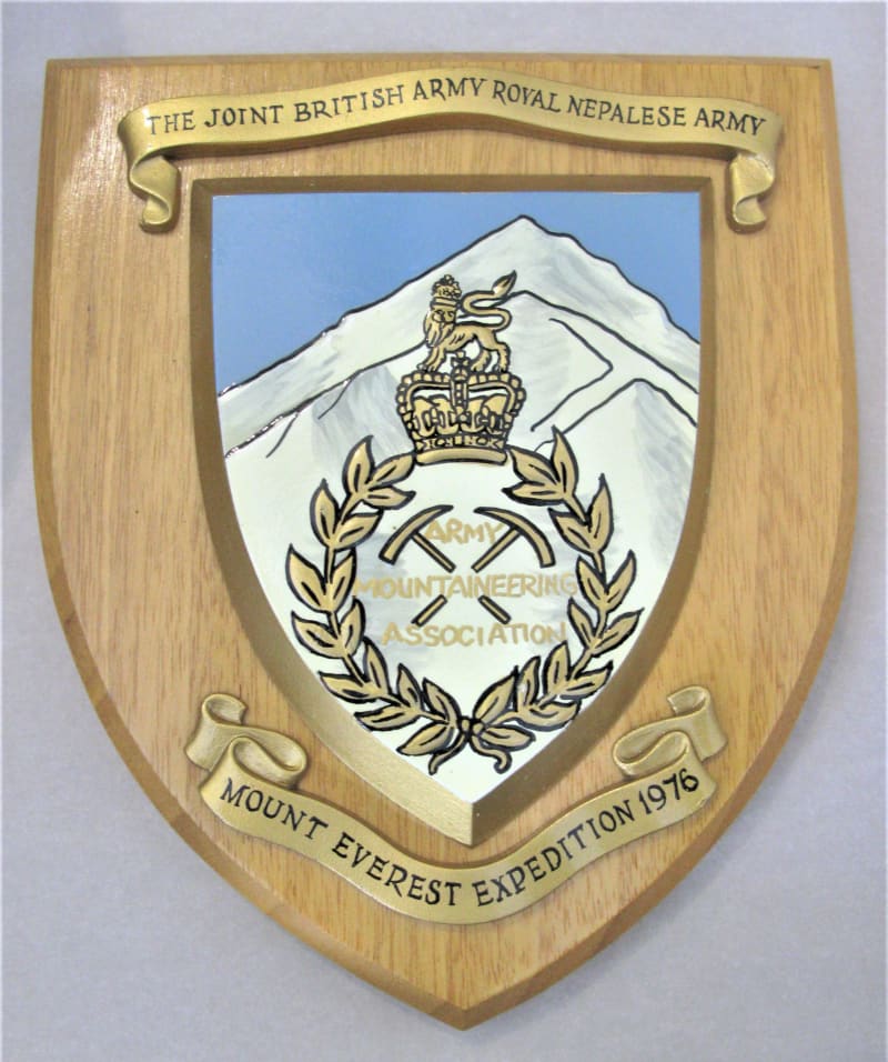 A wooden shield-shaped plaque with golden scrolls on top and bottom reading "Mount Everest Expedition 1976". Shield in the centre depicts a white mountain with badge of the Army Mountaineering Association.