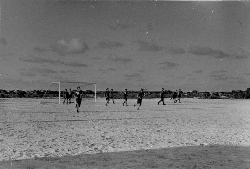 Black and white photograph of a beach with a football goal in the background. Two people stand in the goal and eight more run away from it. All wear t-shirts and shorts