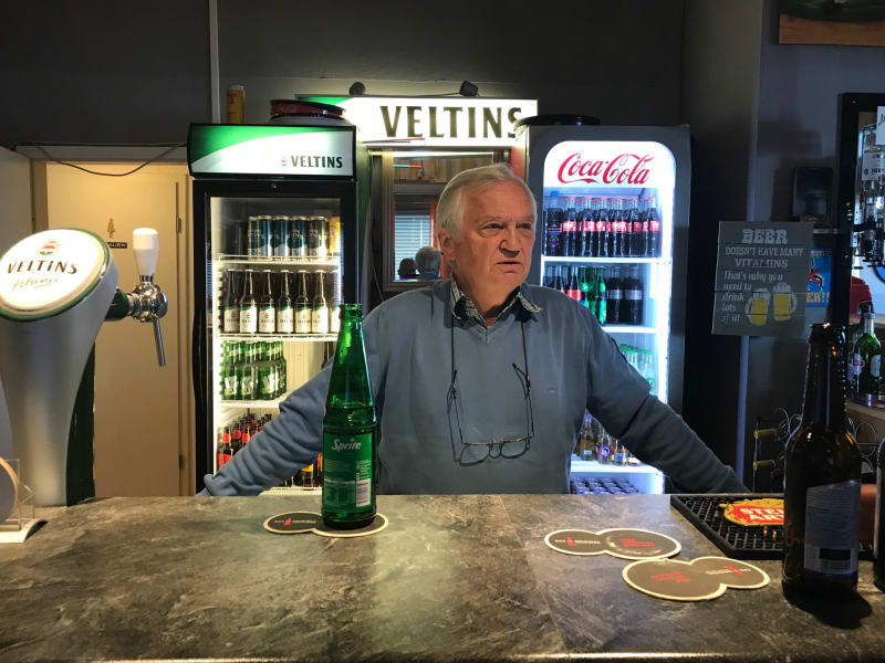 Tom in blue jumper stands behind bar. Glass bottle of sprite and beer mats on bar in front and three fridges with alcohol and coca-cola behind him.