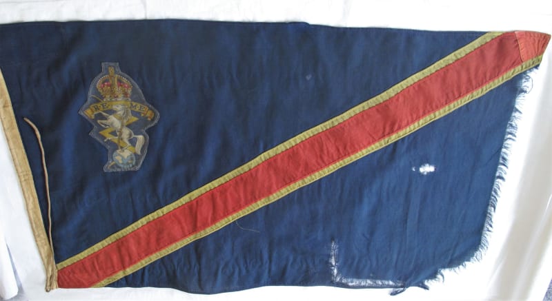 A blue rectangular flag with diagonal red and yellow stripe, REME cap badge in the top left corner, flag is ripped on the right side and torn.