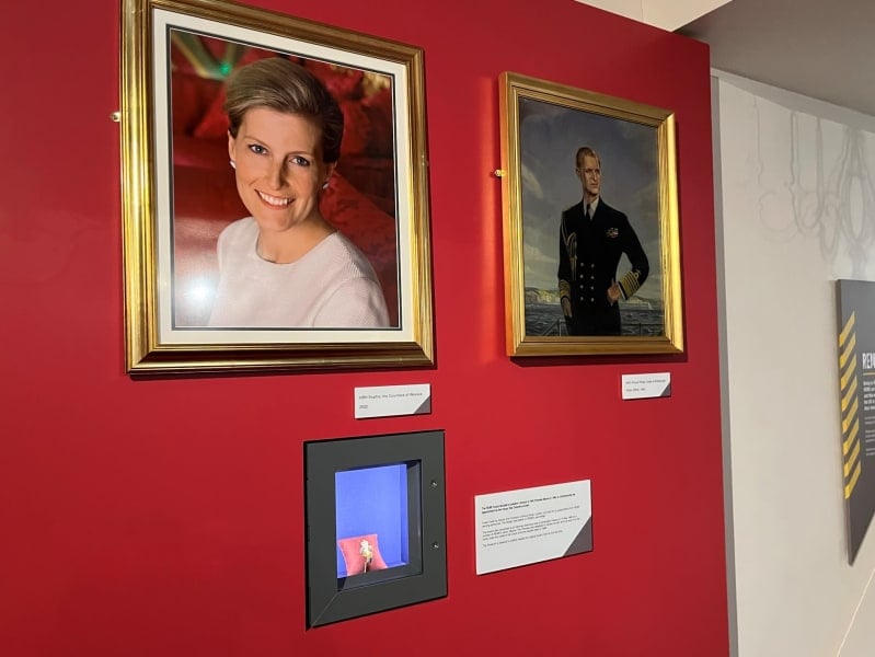 Museum display of two portraits, one of Countess of Wessex and one of Prince Philip, below is a small rectangular display case with brooch sitting on a cushion inside. Red background.